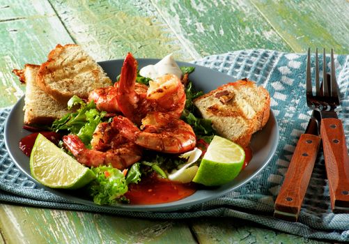 Snack with Big Grilled Shrimps, Fresh Crunchy Greens, Sauces and Roasted Bread Slices on Grey Plate with Fork and Knife closeup on Wooden background