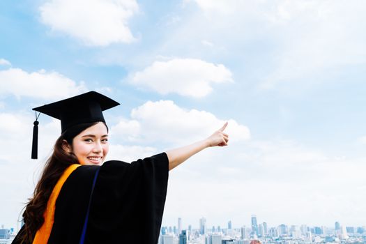 Beautiful asian university or college graduate student woman in graduation academic dress or gown, smiling and pointing at copy space on blue sky background, education or success concept