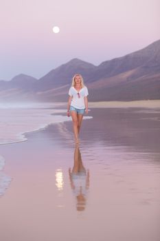 Woman walking on sandy beach in dusk leaving footprints in the sand. Beach, travel, concept. Copy space. Vertical composition.