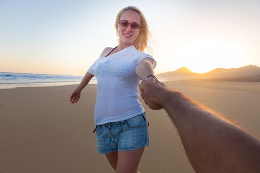 Young romantic couple, holding hands, having fun on perfect deserted beach at sunset. Shot from boyfrieds perspective. Guy looking at her beautiful carefree girlfriend.