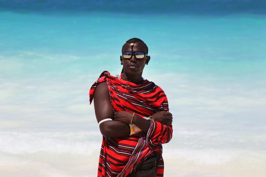 Zanzibar, Tanzania - January 5, 2016: Young massai man with colorful traditional clothes, headdress and self made shoes walking through the water on the Nungwi beach.