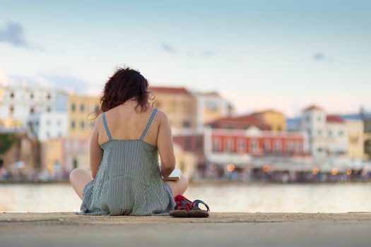 Rear view of woman sitting on a pier near sea port of Chania, Greece, reading book, enjoying vacations.