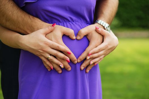 Pregnant Woman and Her Husband holding her hands in a heart shape on her baby bump. Young couple expecting a baby. Waiting for baby. Happy man hugging his pregnant woman.
