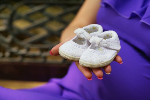 Pregnant woman holding baby shoes on her hand near the belly in happy anticipation