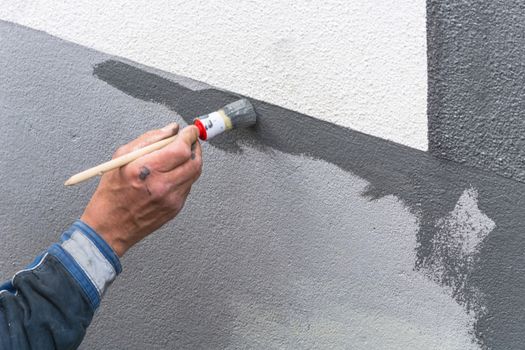 Close-up, arm of a house painter with paint roller in hand painting a house wall with gray paint.
