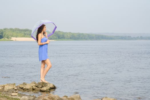 Lonely sad young girl with an umbrella stands on the bank of the river and looks into the distance
