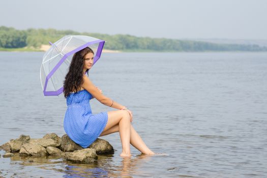 Lonely sad young woman with an umbrella sits on a rock by the river