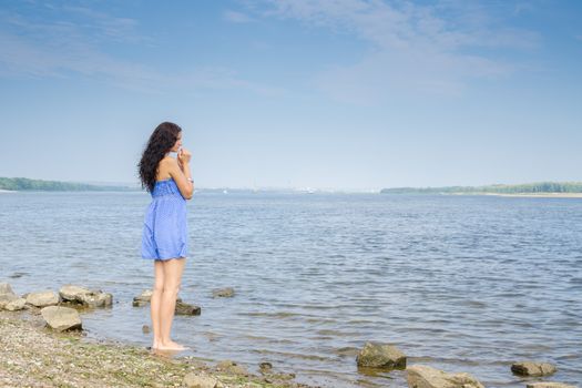Sad young brunette woman in a blue summer dress standing on the river bank