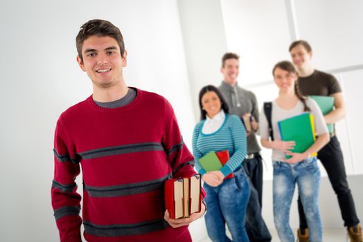 Beautiful smiling young man with book standing in school hall and looking at camera. Happy group of his friends is behind him. 