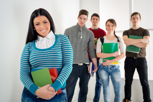 Beautiful smiling student girl with book standing in school hall and looking at camera. Happy group of his friends is behind him. 