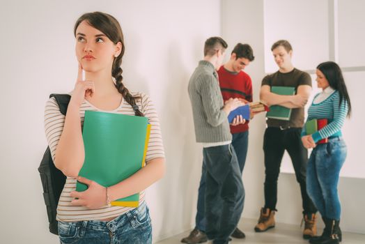 Beautiful pensive student girl standing in school hall and thinking. Happy group of his friends is behind him. 