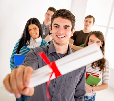 Beautiful successful smiling student with diploma standing in school hall. Happy group of his friends standing is behind him and looking at camera. Selective focus. 