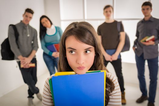 Beautiful student girl standing in school hall and thinking. A happy group of his friends is behind him. 