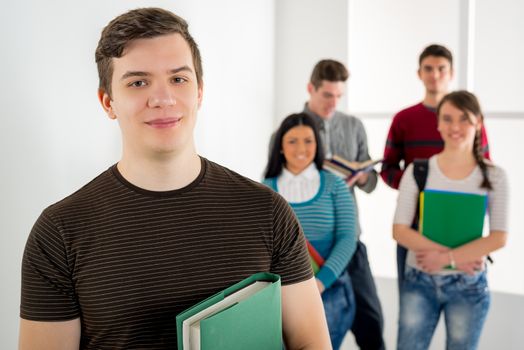 Beautiful smiling young man with book standing in school hall and looking at camera. Happy group of his friends is behind him. 