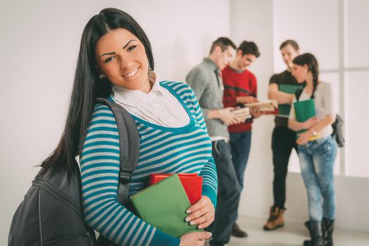 Beautiful smiling student girl with book standing in school hall and looking at camera. Happy group of his friends is behind him. 