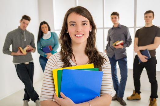 Beautiful smiling student girl standing in school hall and looking at camera. A happy group of his friends is behind him. 