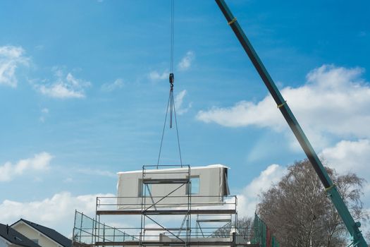 A wall part of a prefabricated house on a crane against blue sky.