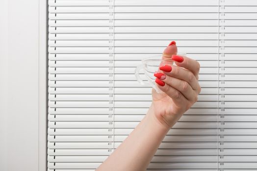 The female hand with beautiful manicure holds a glass cup against a window with blinds