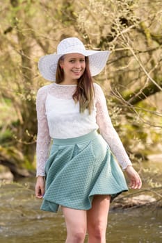 Cheerful fashionable woman in stylish hat and frock posing outdoor in creek. Happy brunette girl with long hair in warm spring day