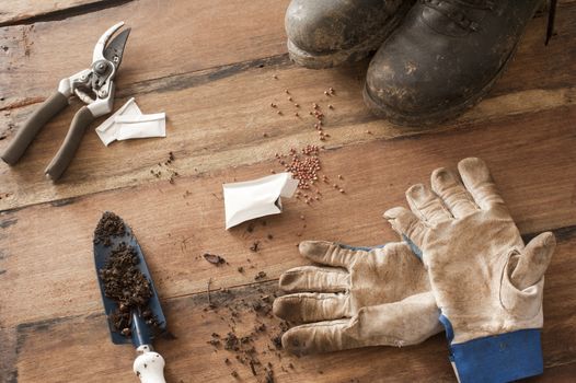 Wood floor with muddy black boots and garden sheers next to open seed packet and gloves by trowel with dirt