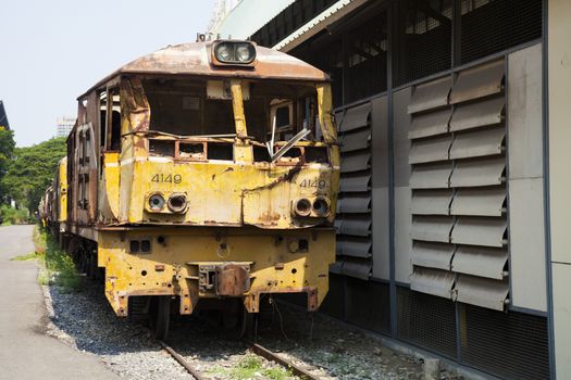 The ancient train has yellow or rust on the old railway near that has rocks and grass and behind has sky tree and buildings.