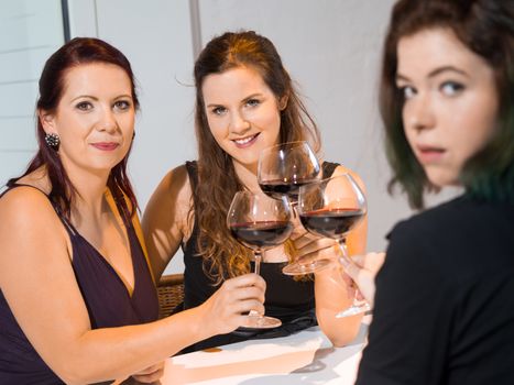 Photo of three female friends toasting with red wine in the kitchen. Focus on middle woman.
