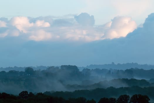 Mist across valley, trees showing in layers and clouds reflecting rising sun.