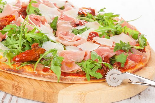 Culinary pizza with prosciutto, dry tomatoes and fresh herbs on wooden cutting board on wooden table. 