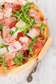 Culinary pizza with prosciutto, dry tomatoes and fresh herbs on wooden cutting board on wooden table. 