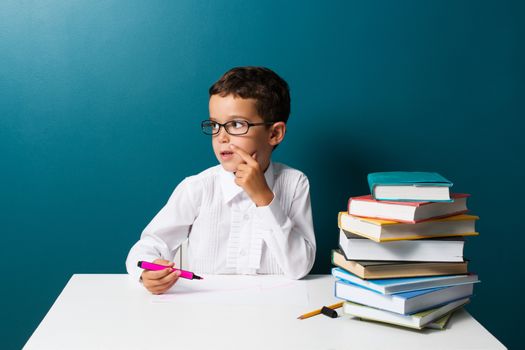 Pensive cute boy with glasses sitting at a table, blue background