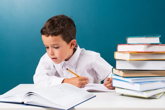 Pensive cute boy with homework sitting at a table, blue background