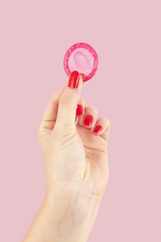 Female hand with red nails holding pink condom isolated on pink background. Safe sex and birth control concept. 