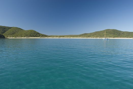 Beautiful Scenic of Single Yacht Sailboat Anchored in Bay near Secluded Tropical Beach on Island Paradise with Rolling Green Hills on Day with Bright Sun and Blue Sky