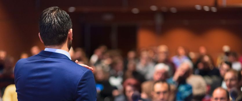 Rear view of speaker giving a talk on corporate Business Conference. Audience at the conference hall. Business and Entrepreneurship event. Panoramic composition.