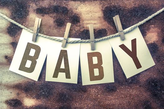 The word "BABY" stamped on cards and pinned to an old piece of twine over a rusted metal background.