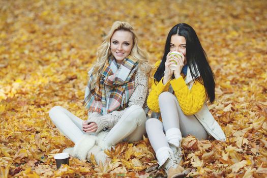 Two cheerful female friends drinking coffee in autumn park