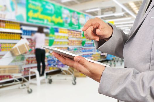 Hand of Businessman use Mobile Tablet in supermarket with Blur Background of Staff arrange product display on shelves in Supermarket or Hypermarket