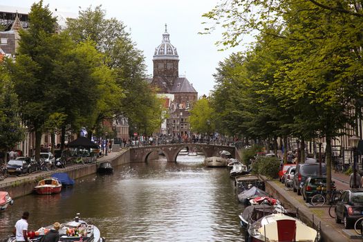 AMSTERDAM, THE NETHERLANDS - AUGUST 16, 2015: View on Saint Nicholas church or St Nicolaas kerk tower from Oudekennissteeg bridge. Street life, Canal, tourists and boat in Amsterdam. Amsterdam is capital of the Netherlands on August 16, 2015.