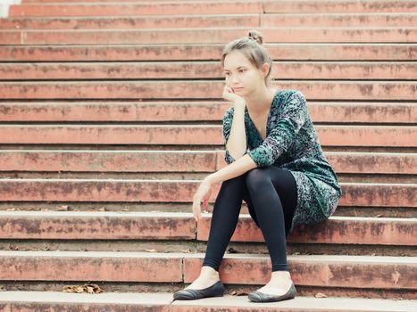 Sad lonely young woman sitting on steps. Portrait of serious girl with facial expression outdoors.