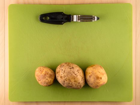 Several three unpeeled potatoes in a row on green plastic board with peeler, simple food preparation illustration, vegetarian dieting, top view still life with copyspace