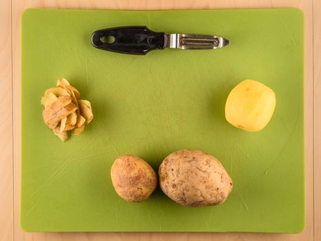 Several unpeeled and peeled potatoes with skins on green plastic board with peeler, simple food preparation illustration, vegetarian dieting, top view still life with copyspace