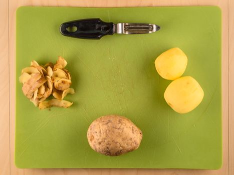 Two unpeeled and one peeled potatoes with skins on green plastic board with peeler, simple food preparation illustration, vegetarian dieting, top view still life with copyspace