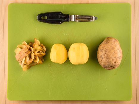 Two unpeeled and one peeled potatoes with skins on green plastic board with peeler, simple food preparation illustration, vegetarian dieting, top view still life with bottom copyspace
