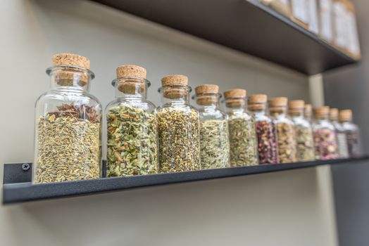 Close-up image with glass bottles filled with different kinds of herbs and plants for tea, displayed on a shelf.