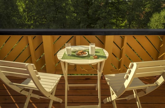 Morning image with a white table and two chairs in a balcony, where it's served the breakfast with milk and cookies.