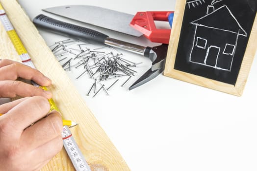 Man's hands  measuring a wooden board, having his tools in the background and a funny sketch of a house on a blackboard. Concept about building your dream house.