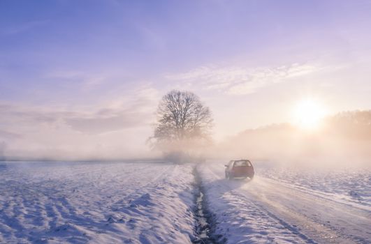 Winter image with the silhouette of a car driving on a snowy country road, through mist and the sunrise light.