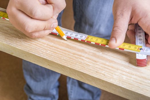 Carpenter is measuring a wooden plank with a measuring tape and making marks with a classic pencil.