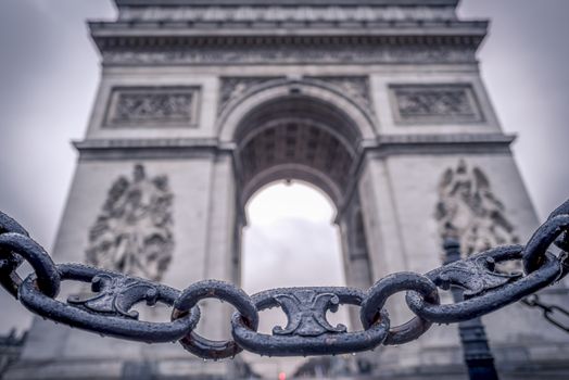 Metaphoric image with chain links sprinkled with rain drops in the foreground and the Arc de triumph on the background, in Paris, France.