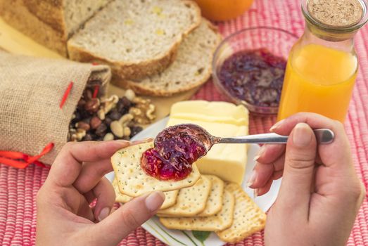 Woman hand holding a cracker with strawberry jam on it and in the background the breakfast table with bread, butter, orange juice and mixed nuts.